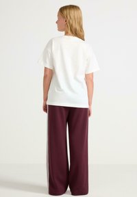 Weißes Baumwoll-T-Shirt mit kurzen Ärmeln, kombiniert mit lockeren, kastanienbraunem Sweatpants, die weiße Seitenstreifen aufweisen. Sichtbare Rückansicht.