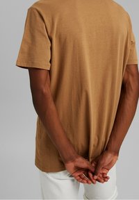 Personne portant une chemise marron à manches courtes et un pantalon blanc, debout les mains jointes dans le dos devant un fond gris uni.