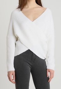 Weißer, weicher Strickpullover mit tiefem V-Ausschnitt und Überkreuz-Design. Rippstruktur und lange Ärmel, kombiniert mit dunkelgrauen Jeans.