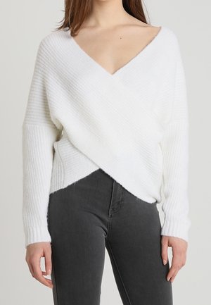 Weißer, weicher Strickpullover mit tiefem V-Ausschnitt und Überkreuz-Design. Rippstruktur und lange Ärmel, kombiniert mit dunkelgrauen Jeans.