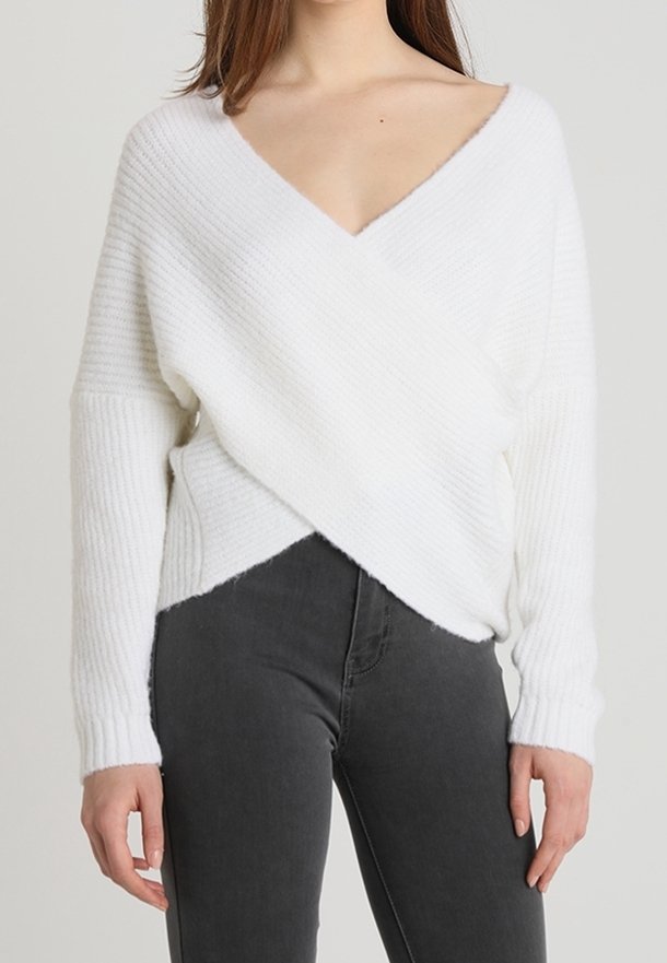 Weißer, weicher Strickpullover mit tiefem V-Ausschnitt und Überkreuz-Design. Rippstruktur und lange Ärmel, kombiniert mit dunkelgrauen Jeans.