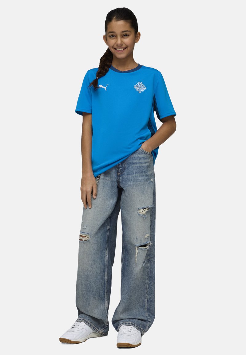 Maglietta sportiva blu a maniche corte in tessuto leggero, abbinata a jeans azzurri strappati e sneakers bianche.