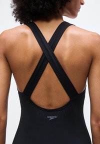 Γυναίκα που φοράει μαύρο μαγιό Speedo με σταυρωτούς ιμάντες που αφήνουν ακάλυπτη την πάνω πλάτη και τους ώμους της, μπροστά σε απλό φωτεινό φόντο.