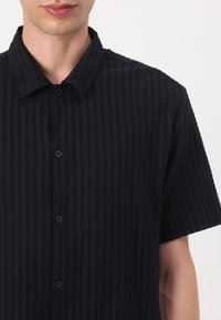 Camicetta nera a maniche corte con motivo a righe texturizzato, chiusura con bottoni anteriori e colletto classico. Tessuto morbido con un design casual.
