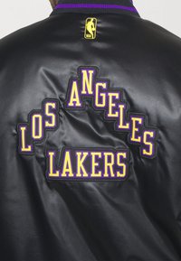 Chaqueta de satén negra con texto bordado "LOS ANGELES LAKERS" en púrpura y amarillo. Cuello acanalado con el logo de la NBA arriba.