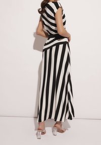 Frau in einem schwarz-weißen, vertikal gestreiften Maxikleid mit einem schwarzen Taillenbund und klaren, hochhackigen Sandalen, stehend vor einer schlichten Wand.