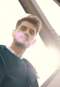 Jonge man in een zwart sweater over een witte blouse, kijkend naar boven met zonlicht en lensflare op de achtergrond.