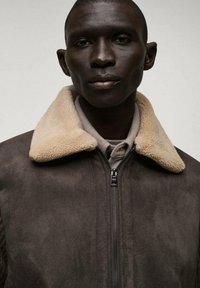 Brun mocka jacka med en beige lammulls-kräm; har en framzipper och en strukturerad design på armarna.