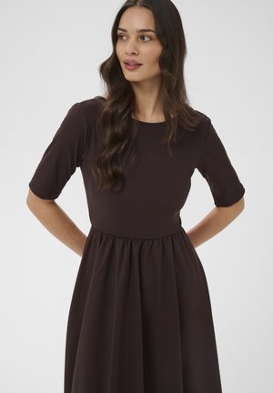 Kobieta z długimi falującymi włosami w ciemnobrązowej sukience z krótkim rękawem, stojąca z rękami za plecami, lekko spoglądająca na bok.