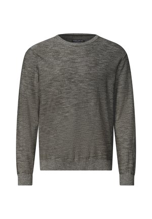 Homme aux cheveux foncés et barbe portant un pull marron texturé et un pantalon beige, légèrement de profil devant un fond clair uni.