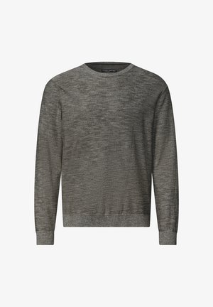 Homme debout avec une main dans la poche, portant un pull marron, un pantalon beige et des baskets blanches sur un fond clair uni.