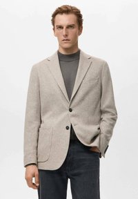 Hellgrauer Blazer aus strukturiertem Stoff, mit zwei Taschen vorne, Revers mit Kerbe und zwei schwarzen Knöpfen. Getragen über einem dunkelgrauen Rollkragenpullover.