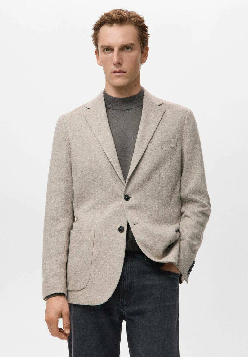 Hellgrauer Blazer aus strukturiertem Stoff, mit zwei Taschen vorne, Revers mit Kerbe und zwei schwarzen Knöpfen. Getragen über einem dunkelgrauen Rollkragenpullover.