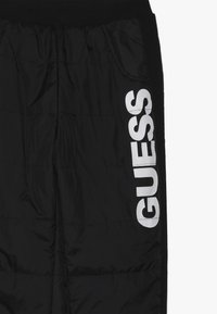 Černé polstrované kalhoty s boční kapsou a velkým bílým logem "GUESS" na levé noze. Měkký materiál s elastickým pasem a manžetami.