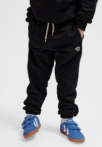 Schwarze Jogginghosen mit elastischen Bündchen, ausgestattet mit einem Kordelzug an der Taille und einem kleinen weißen Logo. Kombiniert mit blauen Klettschuhen.