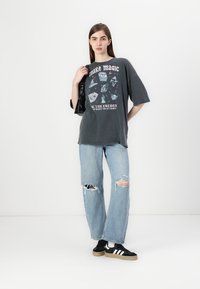 T-shirt graphique surdimensionné gris avec impression multicolore et texte, associé à un jean déchiré bleu clair et des baskets noires.