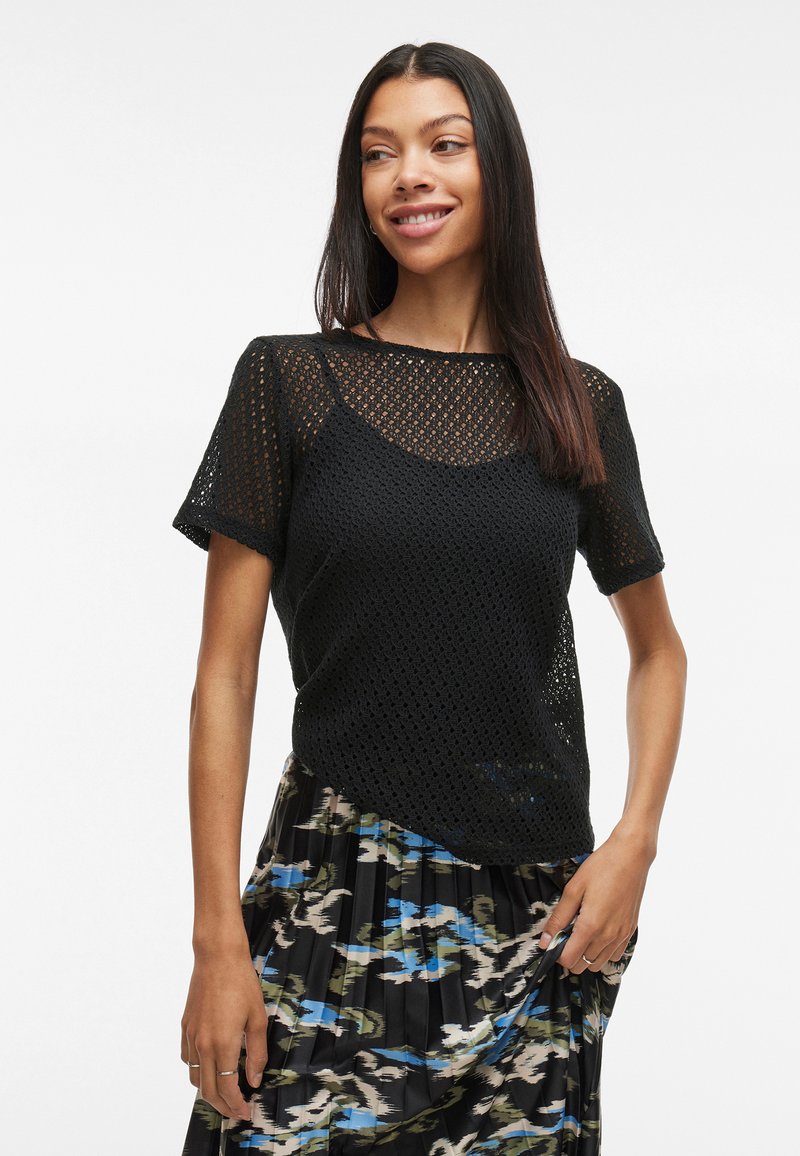 Svart strikket topp med mesh-design, kortermet og rund hals, parret med et mønstrete skjørt i blå og grønne toner.