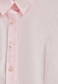 Camicia button-up rosa chiaro con una texture morbida, dotata di bottoni rotondi e una piccola figura bianca ricamata sulla tasca del petto.