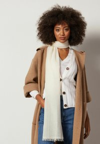 Cremefarvet tørklæde med frynser draperet om halsen, parret med en ribbet hvid sweater og en tanfarvet, overdreven frakke, suppleret med blå jeans.