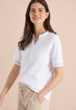 Kobieta o długich brązowych włosach uśmiechająca się, ubrana w biały krótki top z koronkową lamówką i beżowe spodnie ze sznurkiem na neutralnym tle.
