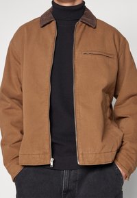 Brun canvasjakke med en kordfløjlskrave, frontlynt og en lille lynlåslomme på brystet. Båret over en sort ribbet turtleneck.