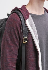 Mörkröd stickad huvtröja med vit inredning, bärs över en grå skjorta. Svart ryggsäksrem med gyllene detaljer synlig. Strukturerad tyg.