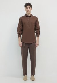 Junger Mann steht mit dem Gesicht nach vorne, trägt ein braunes langärmeliges Hemd mit Knöpfen und passende braune Hose sowie beigefarbene Slipper.