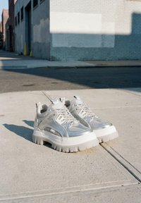 Ασημένια chunky platform sneakers με παχιά λευκή σόλα τοποθετημένα δίπλα-δίπλα σε πεζοδρόμιο από σκυρόδεμα κάτω από έντονο ηλιακό φως.