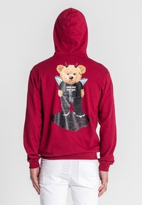 Crvena kapuljača s grafikom medvjeda s rogovima i krilima, koji nosi crnu majicu s natpisom "ANĐELSKO LICE, ĐAVOLJSKA DUŠA", na pozadini oblaka.