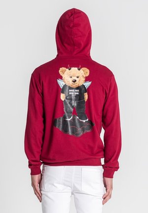 Červená mikina s kapucňou so grafickým motívom medveďa s rohmi a krídlami, oblečeného v čiernom tričku s nápisom "ANJELOVÁ TVÁR, DÉMONSKÁ DUŠA" na pozadí oblakov.