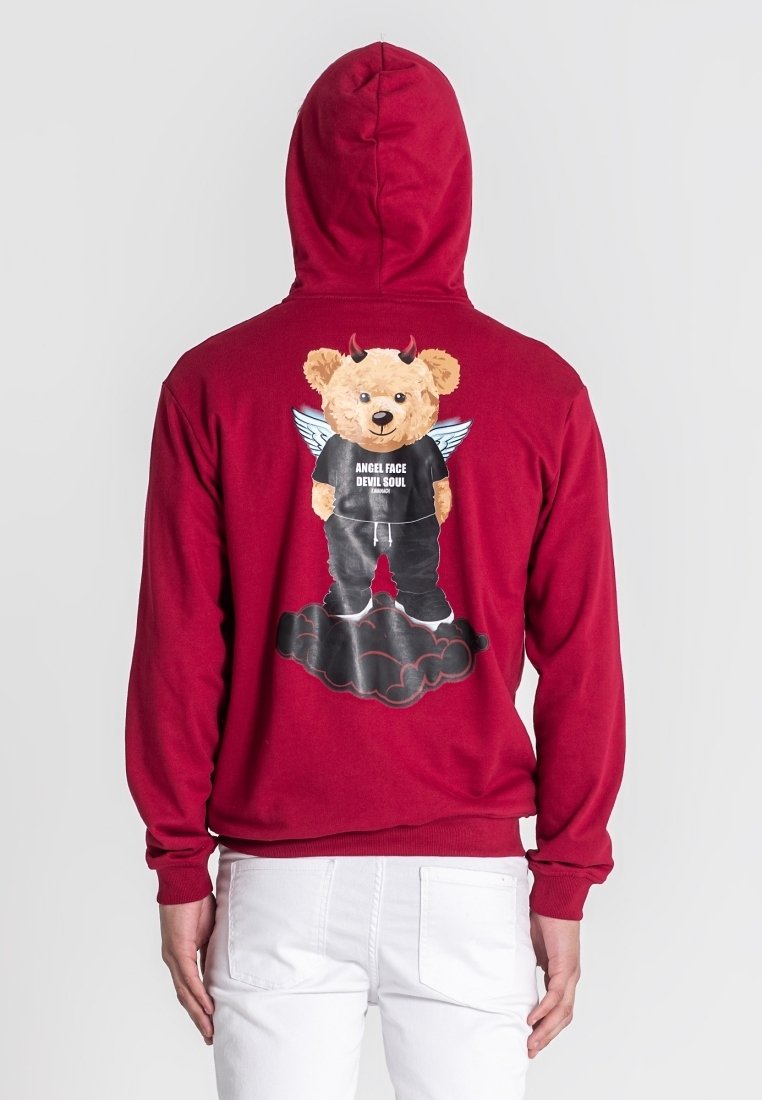 Crvena kapuljača s grafikom medvjeda s rogovima i krilima, koji nosi crnu majicu s natpisom "ANĐELSKO LICE, ĐAVOLJSKA DUŠA", na pozadini oblaka.