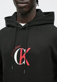 Sweatshirt preta com capuz e cordão, com logótipo da Calvin Klein em vermelho e branco. Tecido macio com textura lisa, vista de perto.