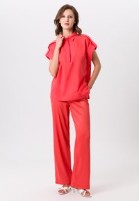 Rood geknoopte shirt met korte mouwen, gecombineerd met bijpassende losse broeken. Gladde textuur met een licht glanzende afwerking. Witte sandalen.