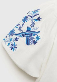 Vit tyg med kort puffärmar har detaljerad broderi i blått och ljusblått av blommor och fåglar på axlarna.