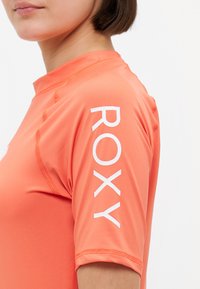 Oransje rash guard med høy krage, korte ermer, flatlock-sømmer, og en dristig hvit "ROXY"-logo på venstre erme. Glatt tekstur.