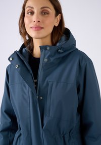 Giacca impermeabile blu navy con cappuccio, chiusure a zip e a pressione, tasca frontale, cordoncino elastico in vita, texture liscia.