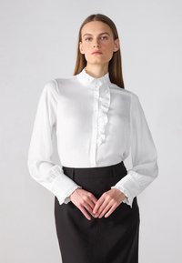 Blouse blanche avec col à volants et fermeture à boutons sur le devant. Les manches longues présentent des poignets à volants. Portée avec une jupe noire, le mannequin pose en position droite.