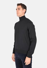 Maglione a collo alto nero con maniche lunghe, polsini a coste e silhouette aderente, realizzato in morbido tessuto lavorato a maglia. Indossato sopra pantaloni in corduroy blu navy.