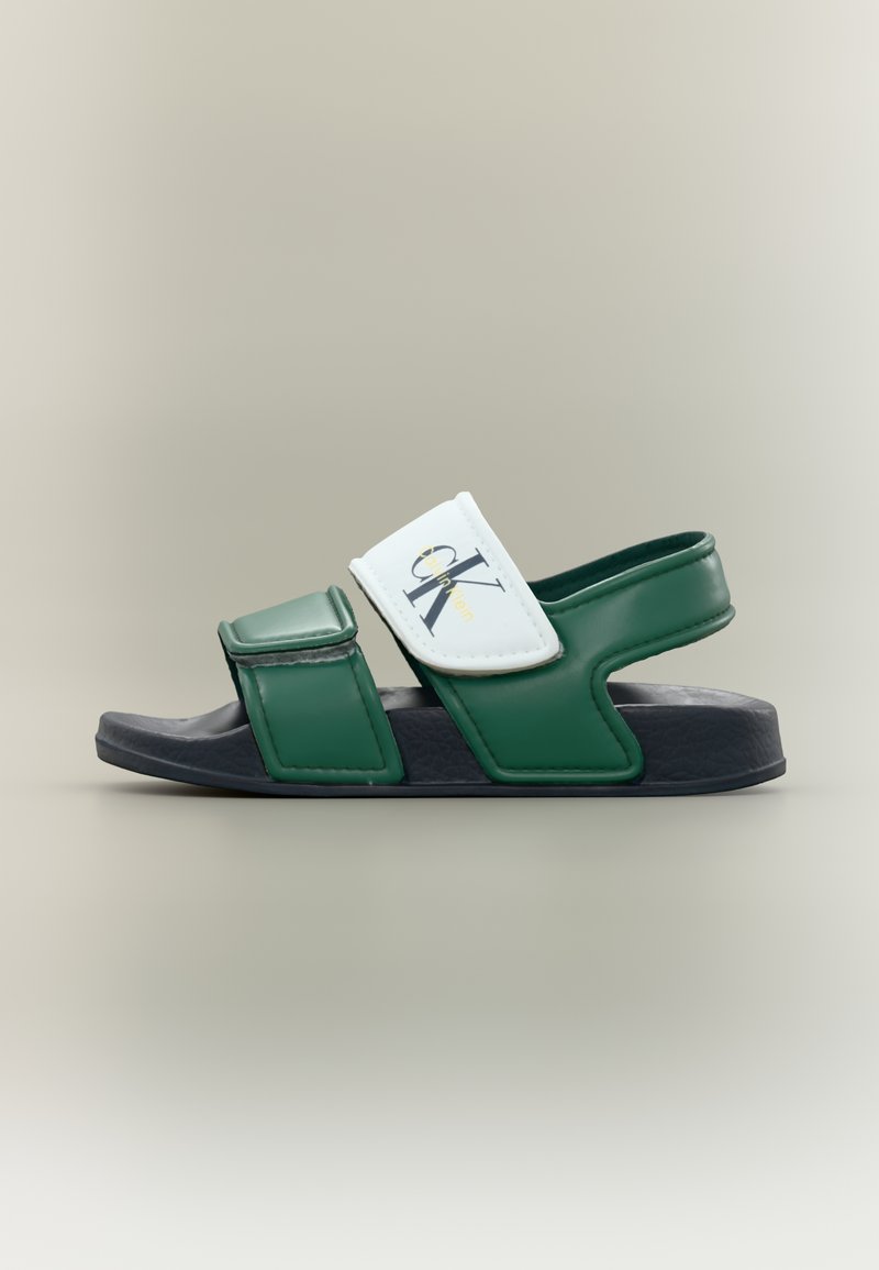Πράσινο και ναυτικό αθλητικό σανδάλι με δύο ρυθμιζόμενα λουράκια, ένα λευκό λουράκι με λογότυπο Calvin Klein, σε ουδέτερο φόντο.