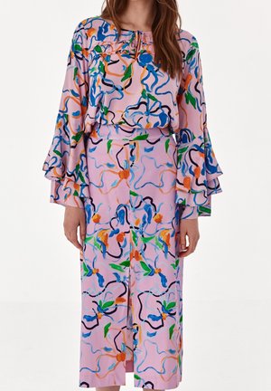 Frau trägt ein langärmeliges, pinkes Kleid mit mehrfarbigen abstrakten Blumenmustern und mehrlagigen Glockenärmeln vor rein weißem Hintergrund.