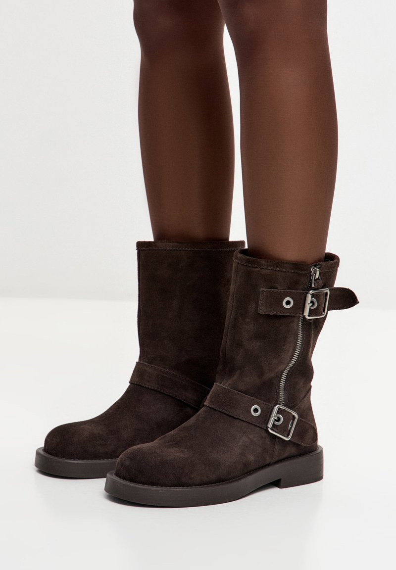 Botine maro din piele întoarsă cu toc jos, închidere cu fermoar lateral și o curea cu accesorii argintii. Textură netedă și design robust.