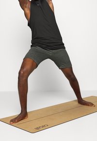 Débardeur noir sans manches, shorts gris foncé, debout sur un tapis de yoga beige avec des lignes imprimées, affichant une posture athlétique et pieds nus.