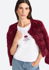 Burgunderfarbene, texturierte Kunstfelljacke über einem weißen T-Shirt mit pinkem Lippenmotiv und dem Text "CHERRY ELEGANCE". Helle blaue Jeans.