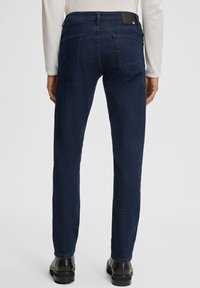 Personne portant un jean droit bleu marine, un haut blanc à manches longues et des bottes noires, vu de dos sur un fond uni.