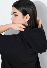 Felpa nera con cappuccio realizzata in un tessuto leggero e texturizzato. La mano della modella tira il cappuccio, evidenziando il suo design strutturato e lo stile minimalista.