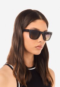 Černé sluneční brýle se čtvercovým tvarem, hnědými tónovanými čočkami a matným povrchem. Na stranicích je zlaté logo a brýle mají minimalistický design.