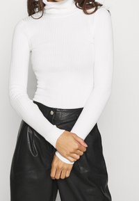 Vrouw die een getailleerde witte geribbelde coltrui en een zwarte leren broek met hoge taille draagt, met haar handen gevouwen ter hoogte van haar taille.