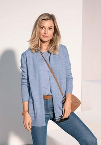 Hellblaues Cardigan mit langen Ärmeln, gerippten Bündchen, offener Vorderseite, kombiniert mit einem blauen Oberteil und dunklen Jeans. Die Accessoires bestehen aus einem hellbraunen Gürtel und einer Handtasche.