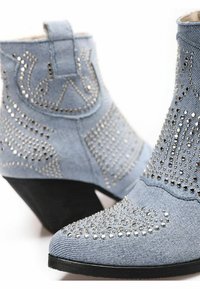 Stivali ankle in denim con punta appuntita, colore azzurro chiaro, borchie decorative in argento e tacco a spillo. Tessuto testurizzato e design con cuciture dettagliate.
