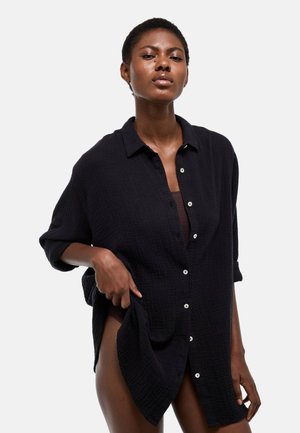 Femme noire portant une chemise noire texturée boutonnée par-dessus des sous-vêtements marron, posant avec une main soulevant la chemise sur un côté.